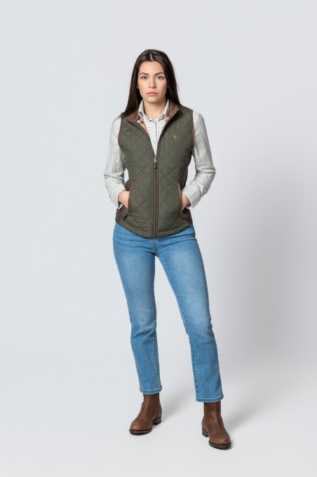 Mulher com colete verde militar matelassê, camisa branca quadriculada, jeans azuis e botins castanhos