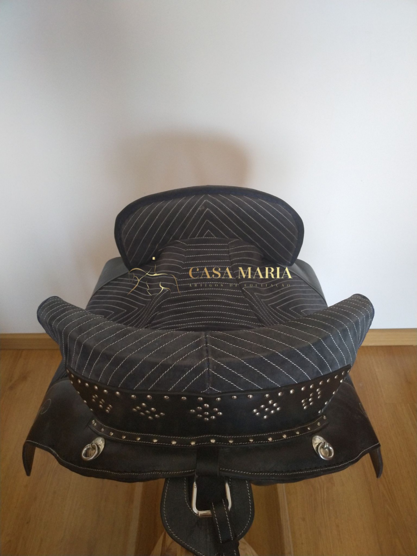 Sela de equitação preta com costura branca e detalhes metálicos sobre chão de madeira