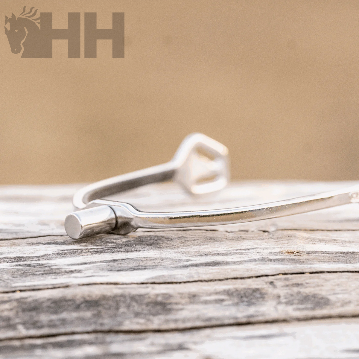 Espora metálica prateada sobre madeira com logotipo HH e cavalo no fundo