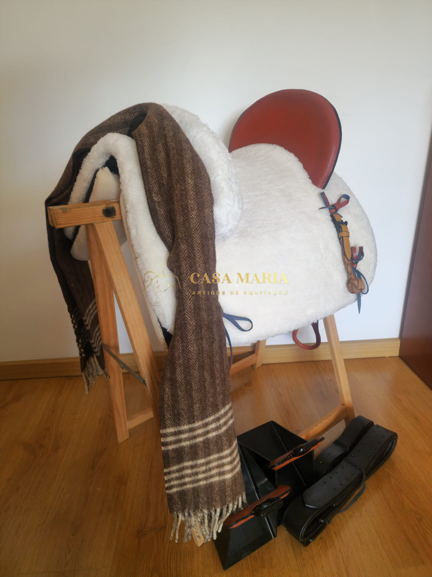 Sela de equitação branca com aba castanha em apoio de madeira, manta castanha com riscas e estribos no chão