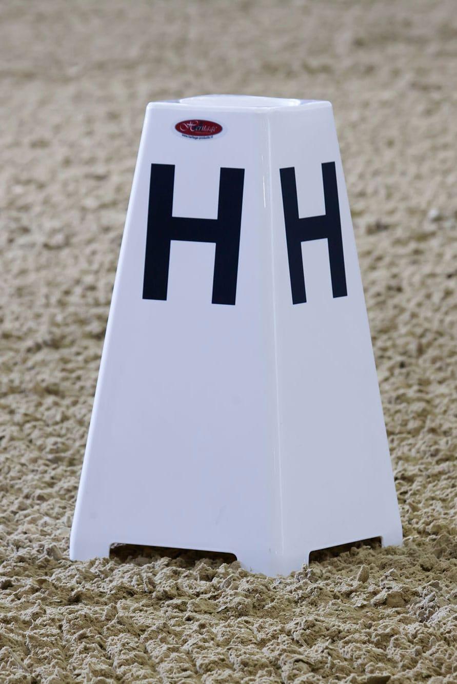 Marcador ou cone plástico branco com letra 'H' preto em piso de areia