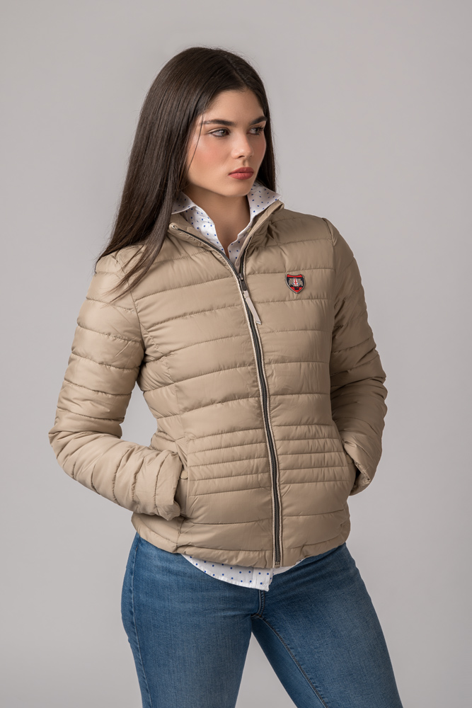 Casaco acolchoado bege com fecho cinzento e emblema no peito, combinado com camisa branca e jeans azuis.