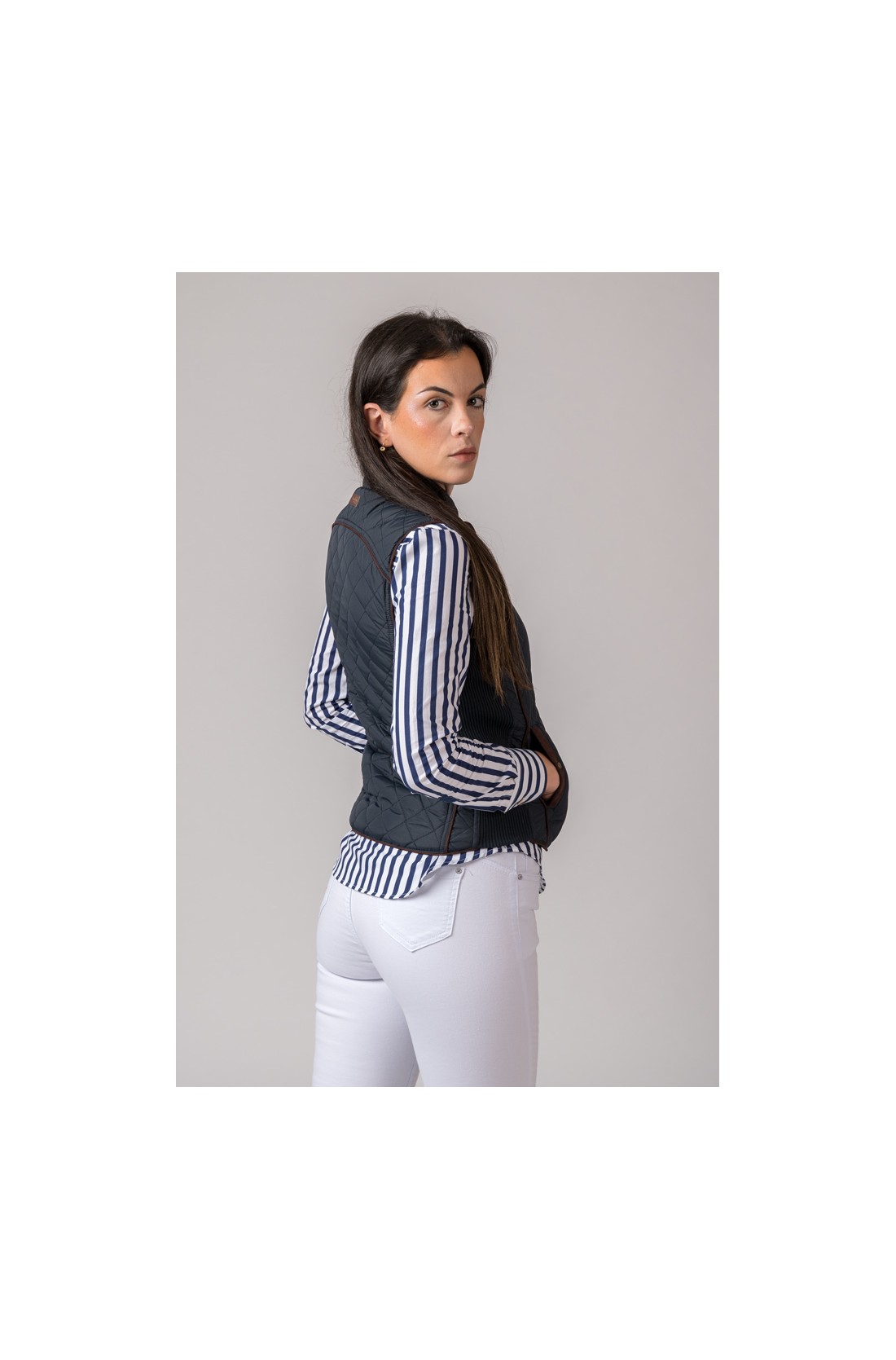 Mulher de costas vestindo colete preto acolchoado, camisa de riscas azuis e calças brancas