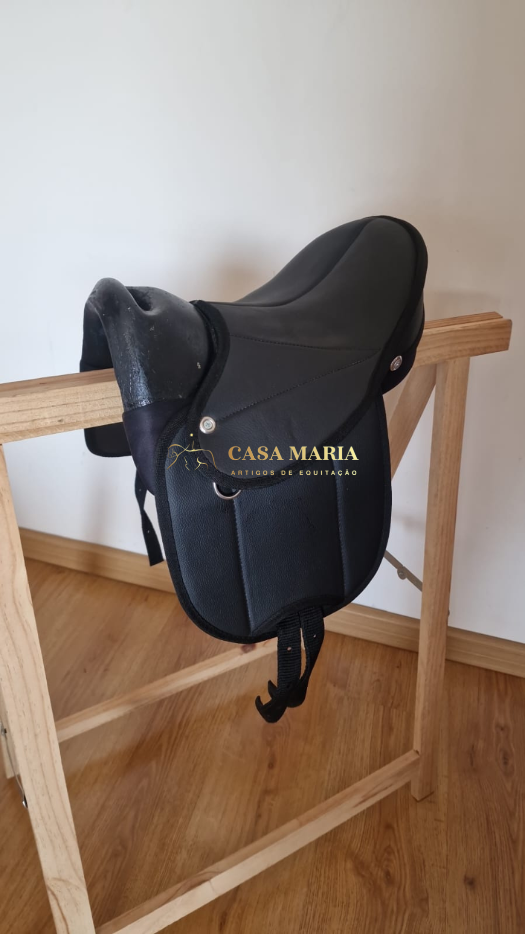 Sela de equitação preta feita de material sintético sobre suporte de madeira com texto CASA MARIA ARTIGOS DE EQUITAÇÃO
