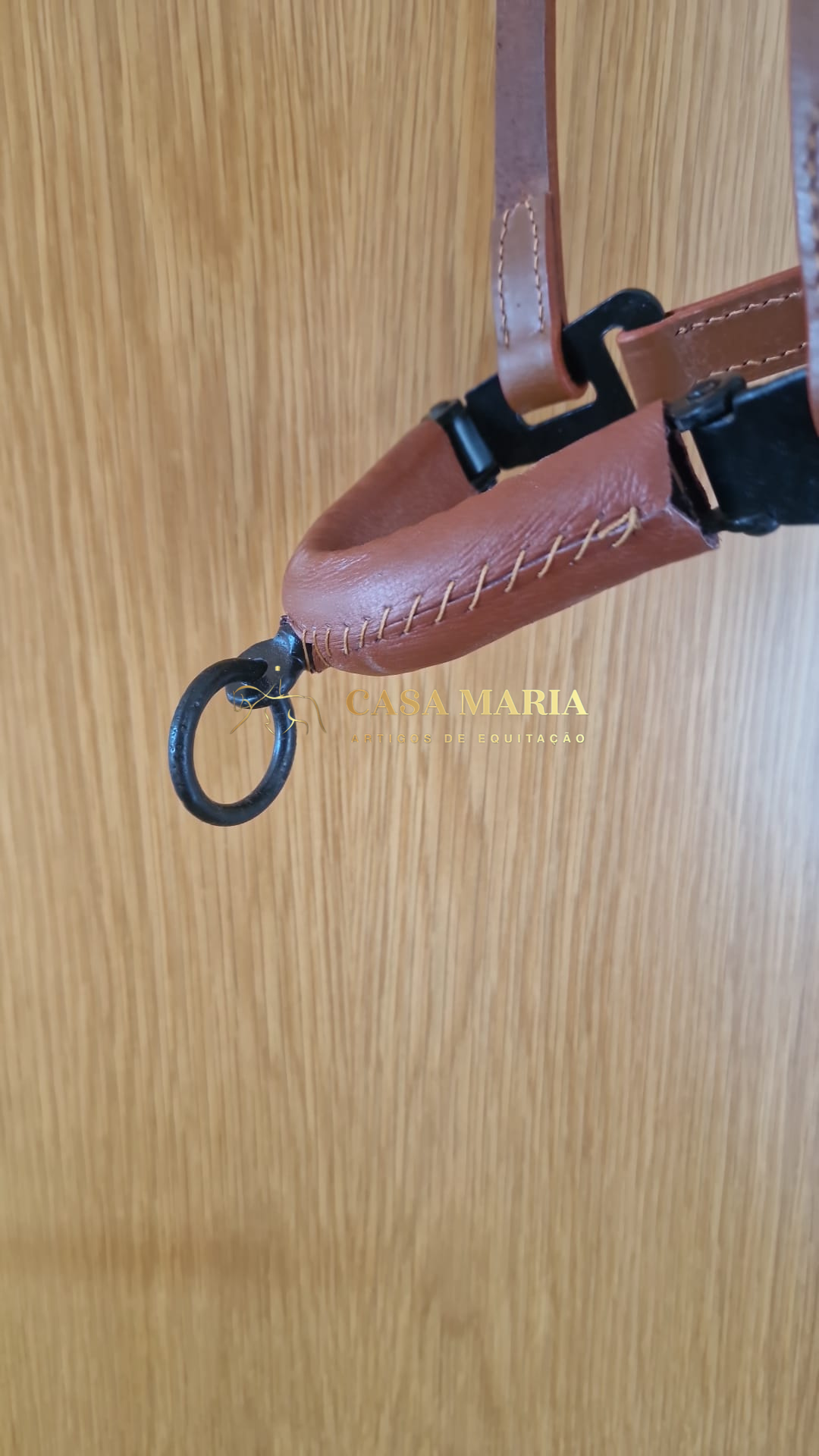 Detalhe de produto de equitação em couro castanho com argola metálica preta e texto CASA MARIA