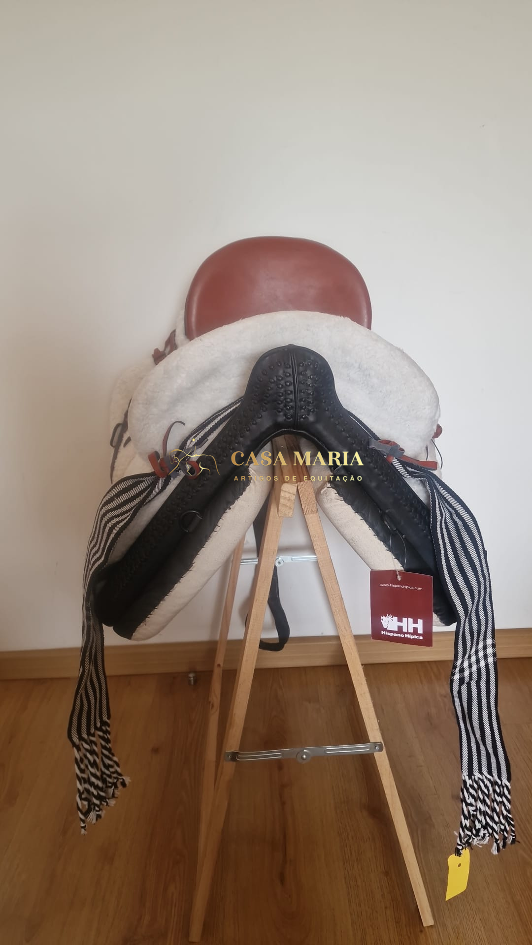 Sela de equitação em couro castanho e preto com acolchoamento branco e correias têxteis penduradas, em suporte de madeira