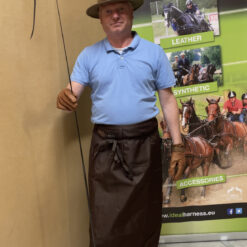 Homem com avental preto e chapéu segurando rédea em frente a banner publicitário de equipamentos para cavalos.