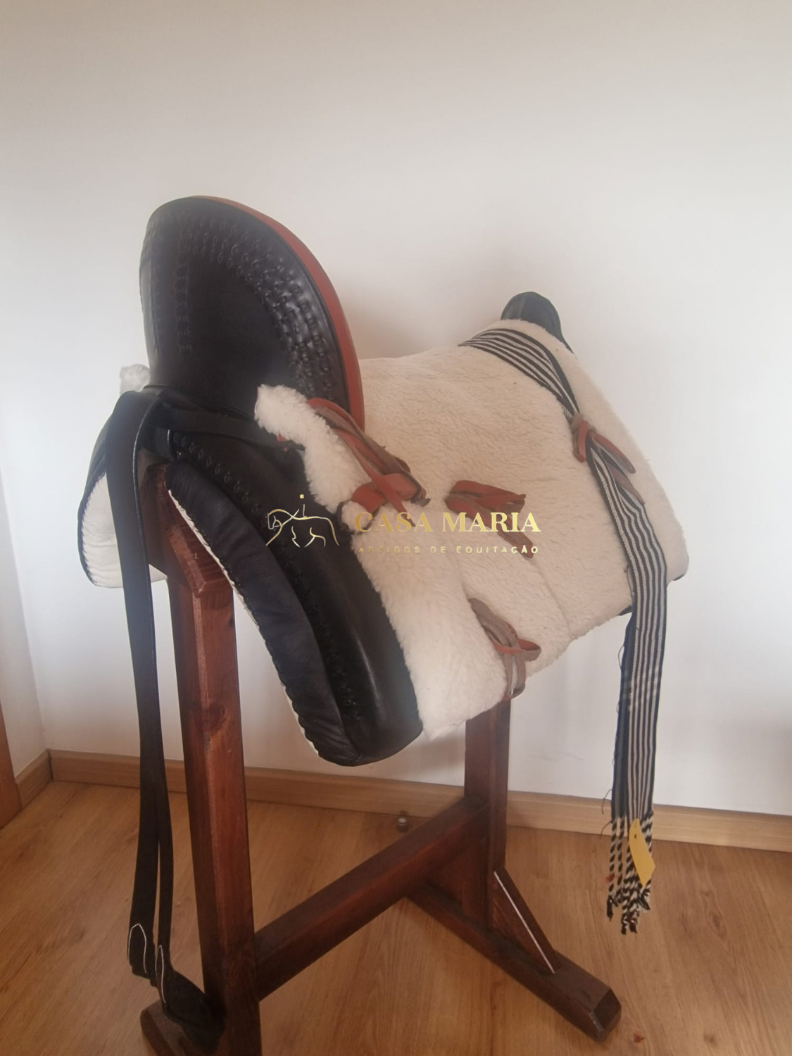 Sela preta acolchoada com lã branca e fita preta e branca, no suporte de madeira, com texto CASA MARIA ARTIGOS DE EQUITAÇÃO