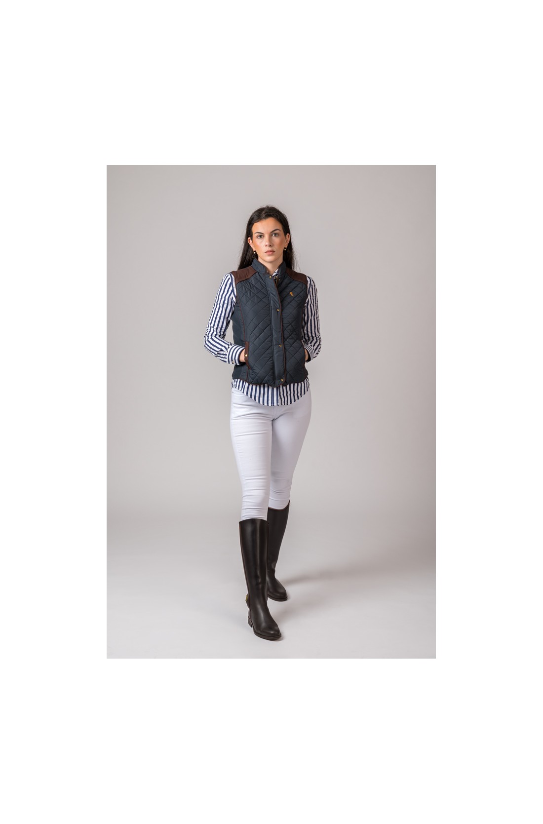 Mulher com colete acolchoado preto, camisa riscada azul e branca, calças brancas e botas pretas numa parede cinza.