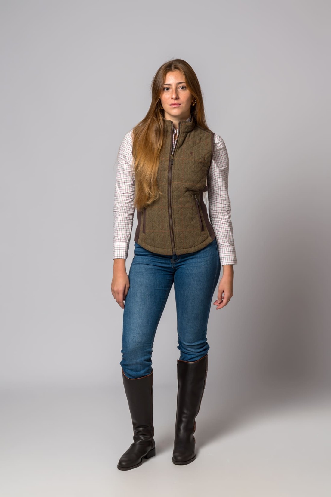 Mulher com colete matelassê, camisa quadriculada, jeans e botas pretas