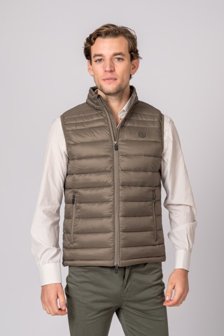 Homem a usar colete acolchoado castanho com fecho frontal e bolsos, camisa bege e calças verdes
