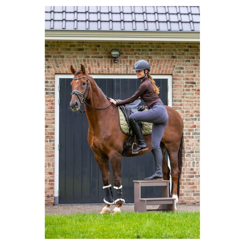 Mulher a montar cavalo castanho com equipamento de equitação, junto a porta preta numa parede de tijolos.