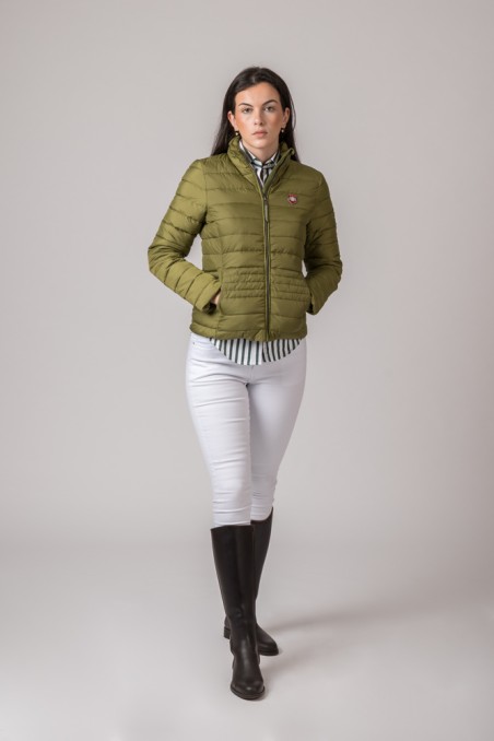 Mulher a usar casaco acolchoado verde oliva com fecho de correr, camisa às riscas, calças brancas e botas pretas.