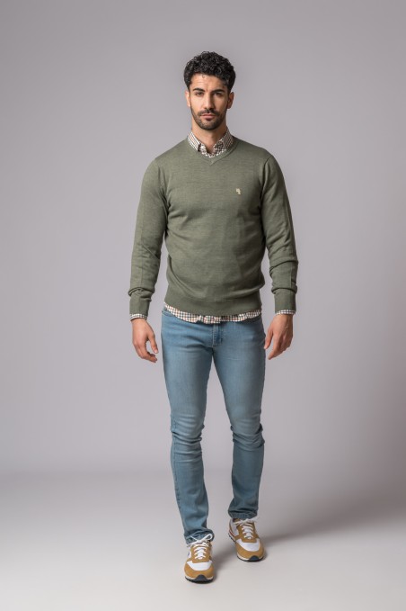 Homem com camisola verde, camisa xadrez, jeans azuis e ténis bege em fundo cinza claro