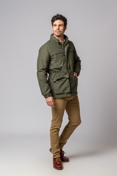 Homem com casaco verde-oliva e calças castanhas em fundo cinzento