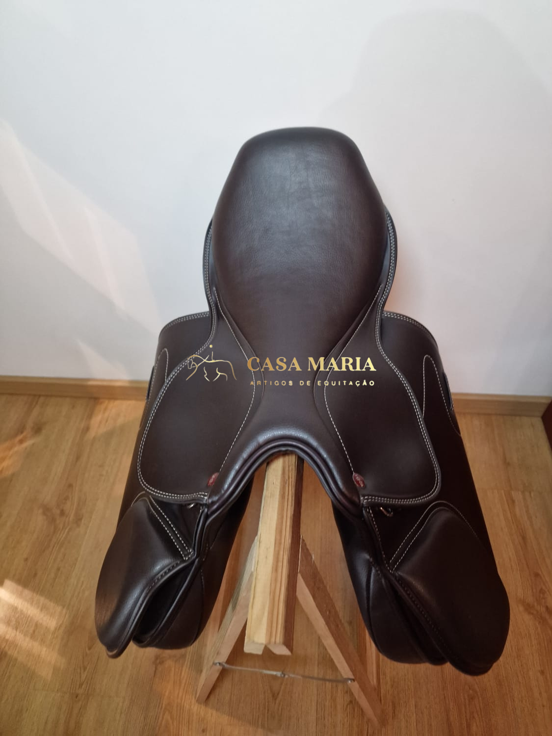 Sela de equitação castanha escura com costuras brancas sobre suporte de madeira