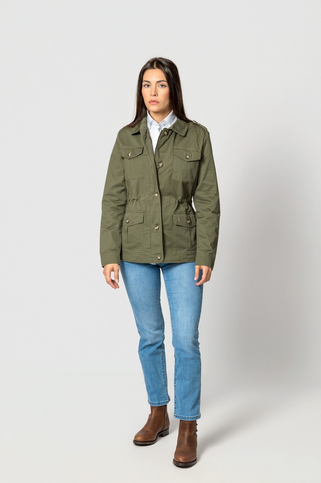 Mulher com casaco verde, jeans e botas em fundo branco.