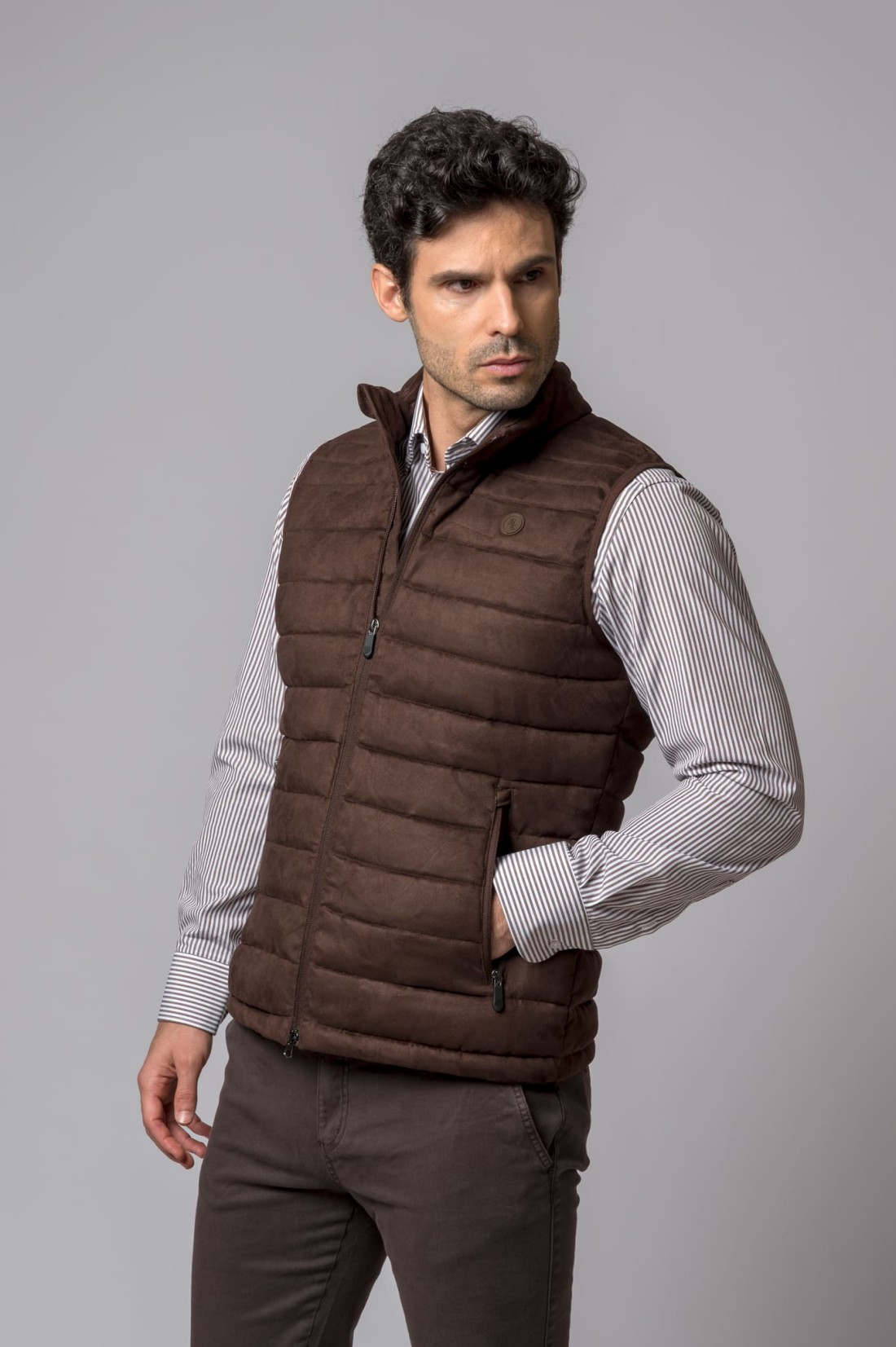 Homem com colete acolchoado castanho e camisa riscada em fundo cinza