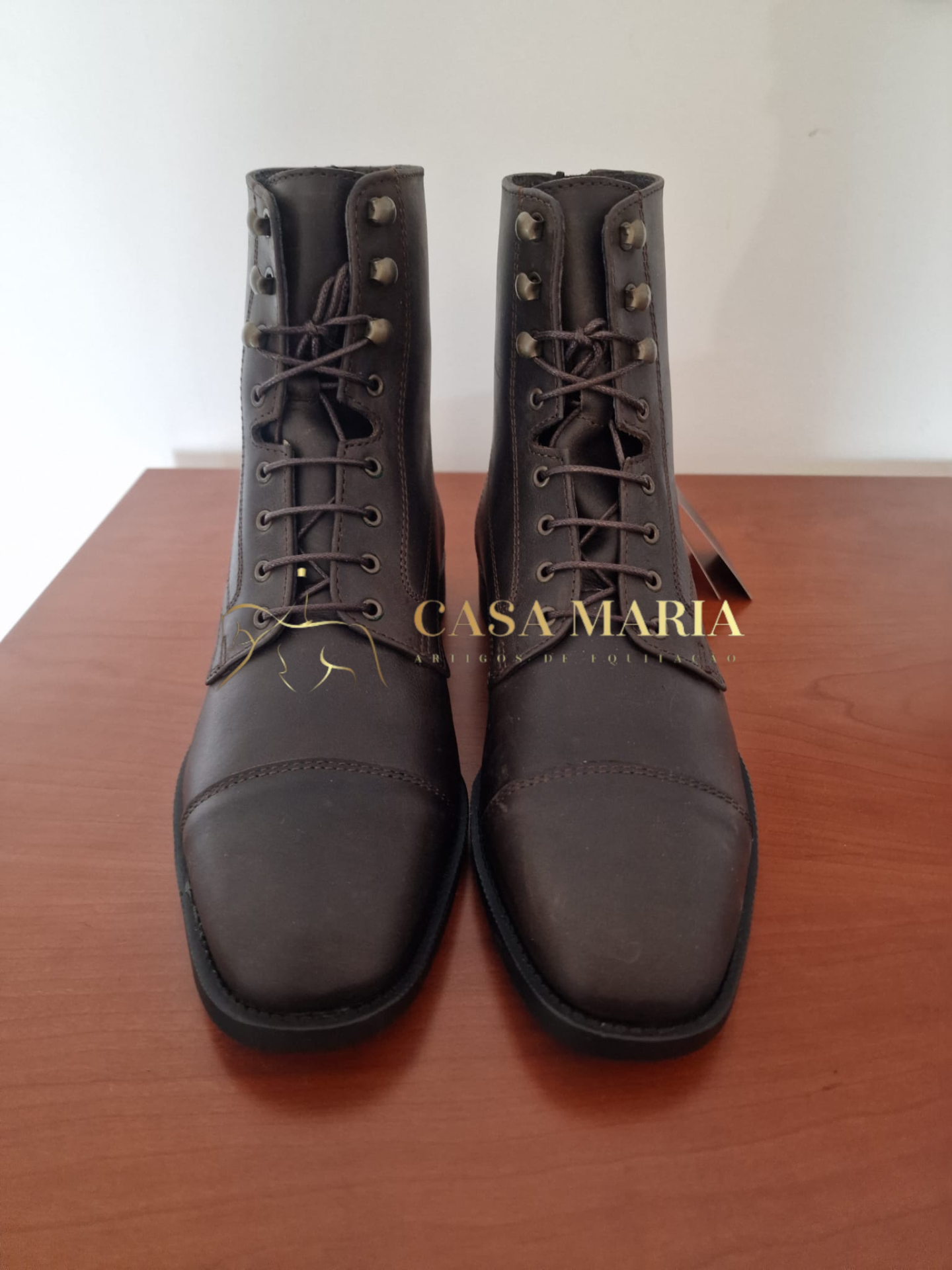 Botas castanho-escuras em couro com atacadores e ilhóses metálicos, sobre mesa de madeira