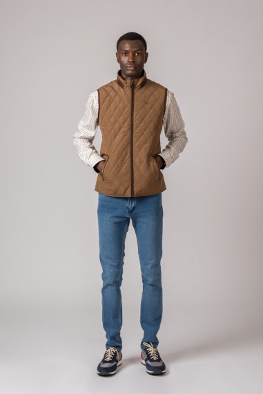 Homem com colete acolchoado castanho, camisa às riscas branca e castanha, jeans azuis e ténis azuis