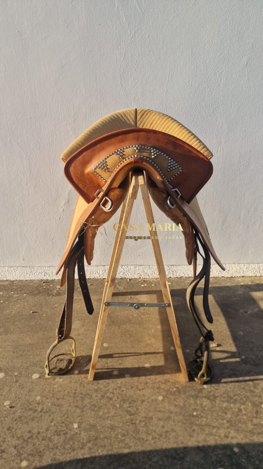 Sela de couro castanho com tachas e estribos pendurados em suporte de madeira