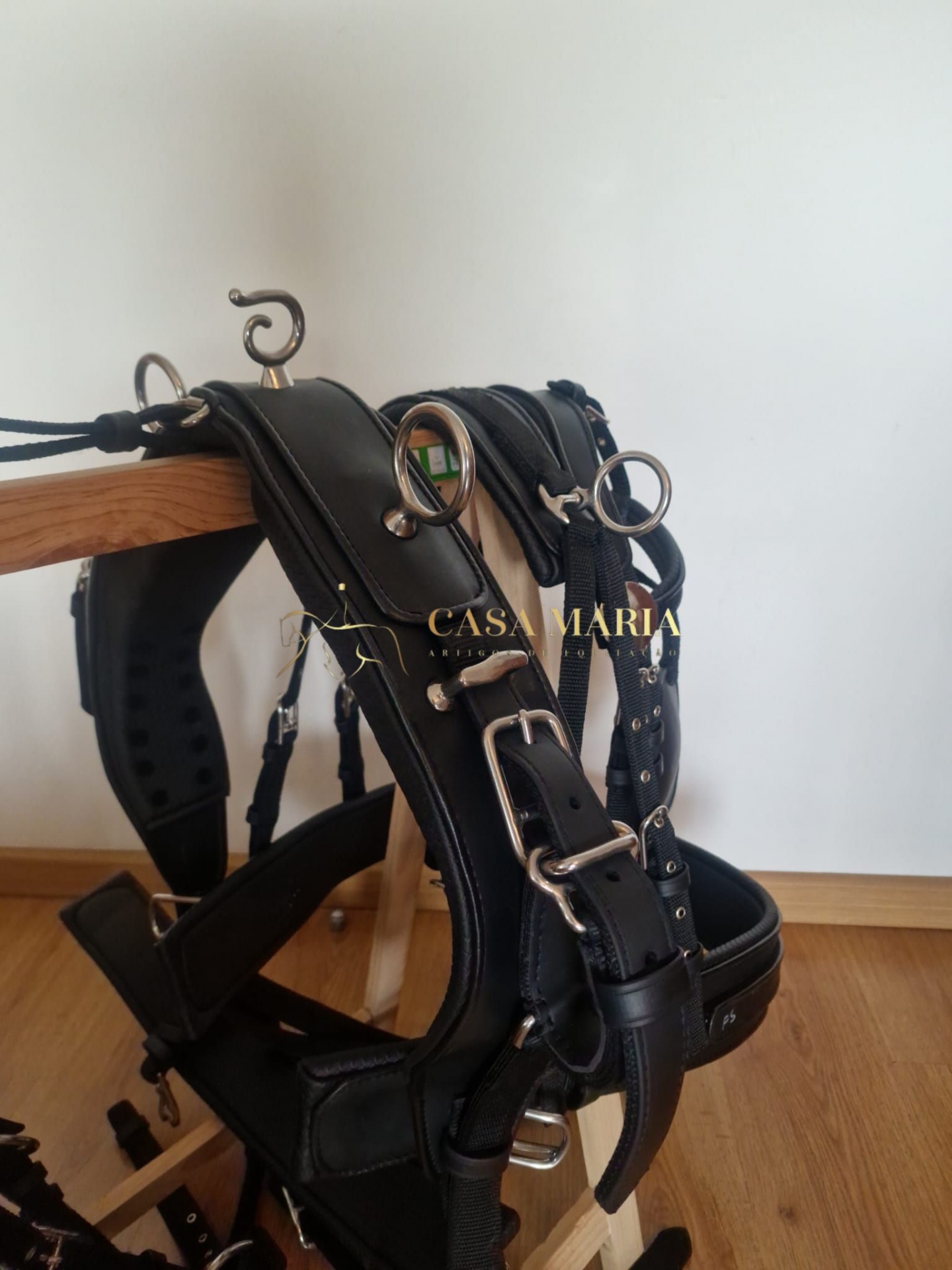 Arnês de couro preto com fivelas metálicas prateadas, pendurado num suporte de madeira
