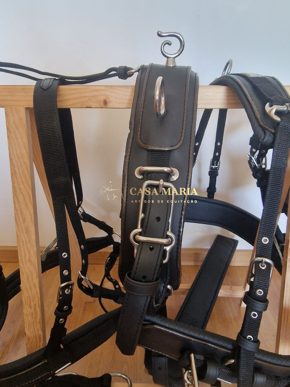 Arnês de equitação preto em couro e nylon com fivelas metálicas pendurado numa estrutura em madeira clara