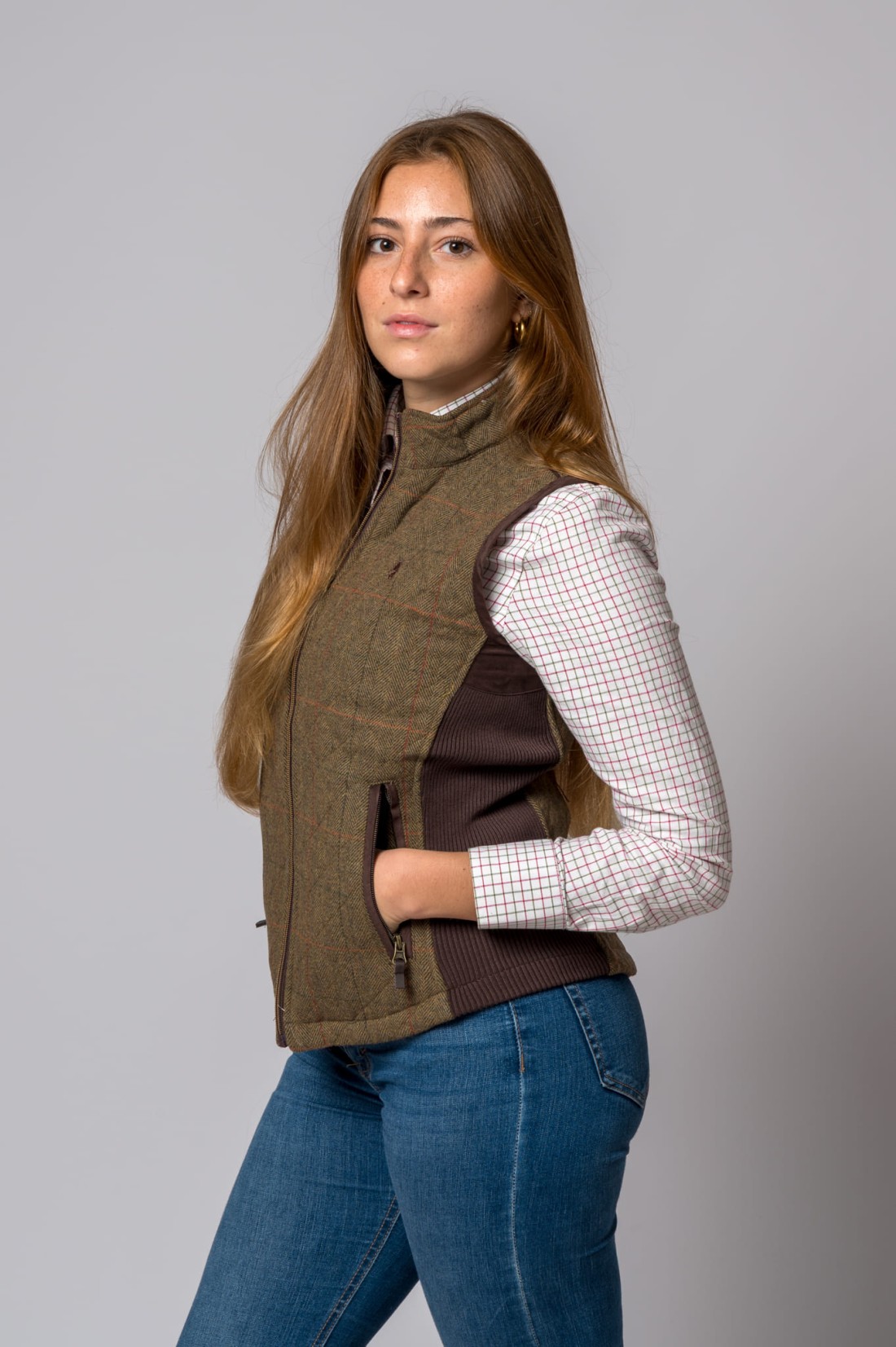 Mulher com colete castanho em xadrez, camisa xadrez branca e jeans azul num fundo cinzento