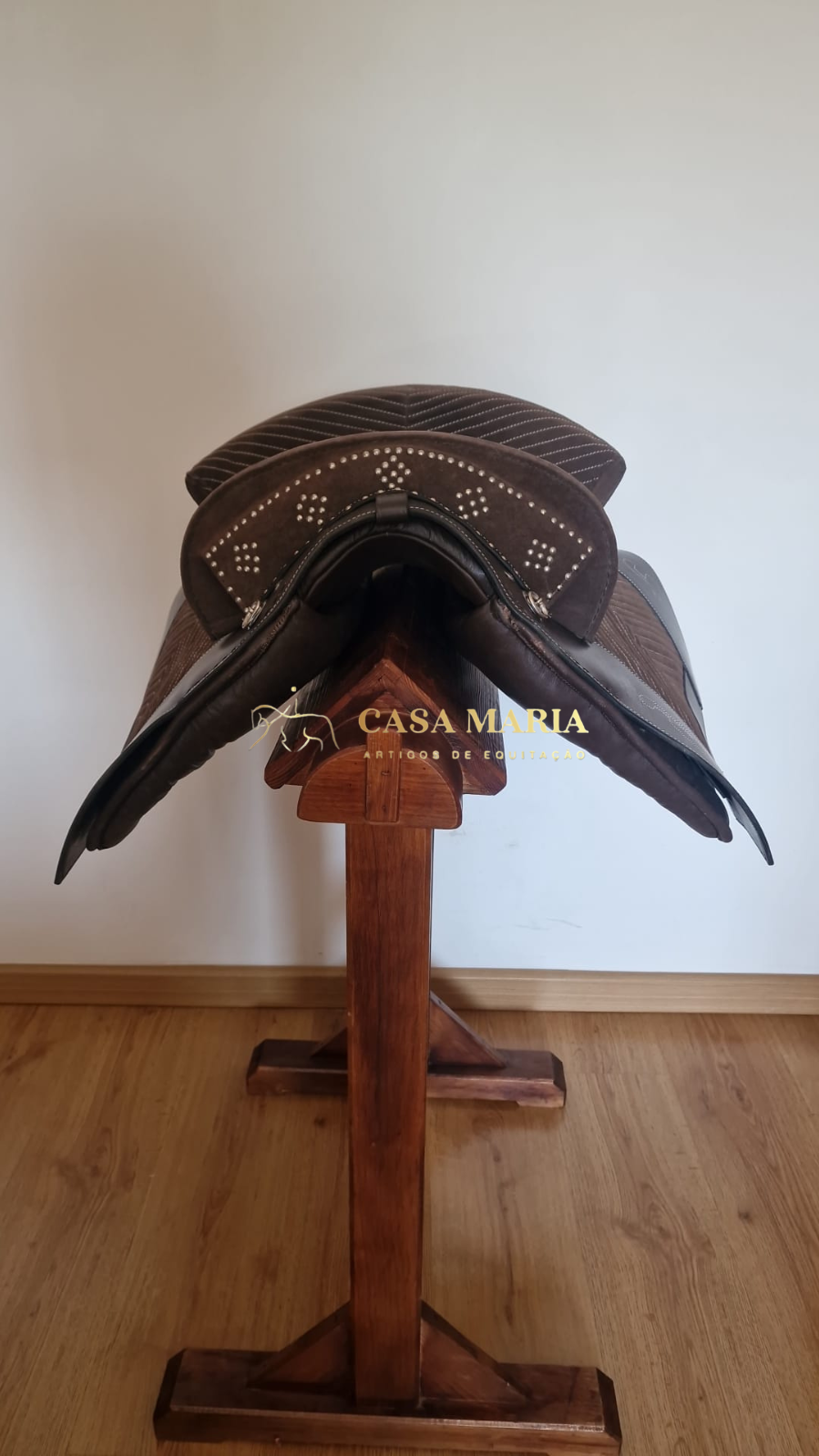 Sela de equitação castanha com padrão decorativo, sobre suporte de madeira e chão de madeira