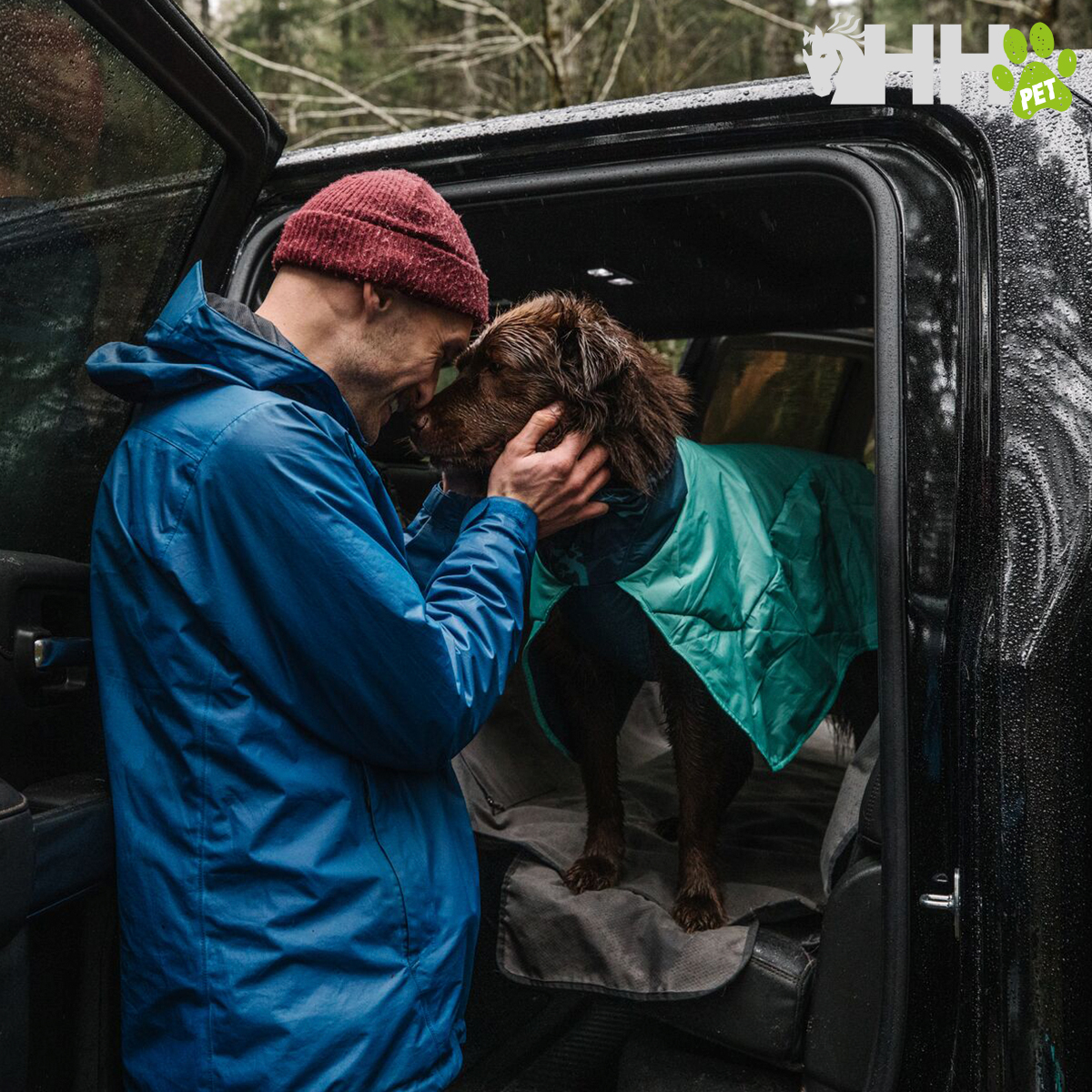 Homem com casaco azul e gorro vermelho e cão com casaco verde num veículo preto em floresta