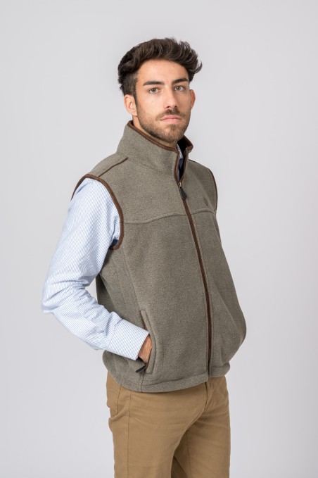 Homem com colete cinza de tecido polar e fecho castanho, camisa azul clara e calças castanhas sobre fundo cinza claro.