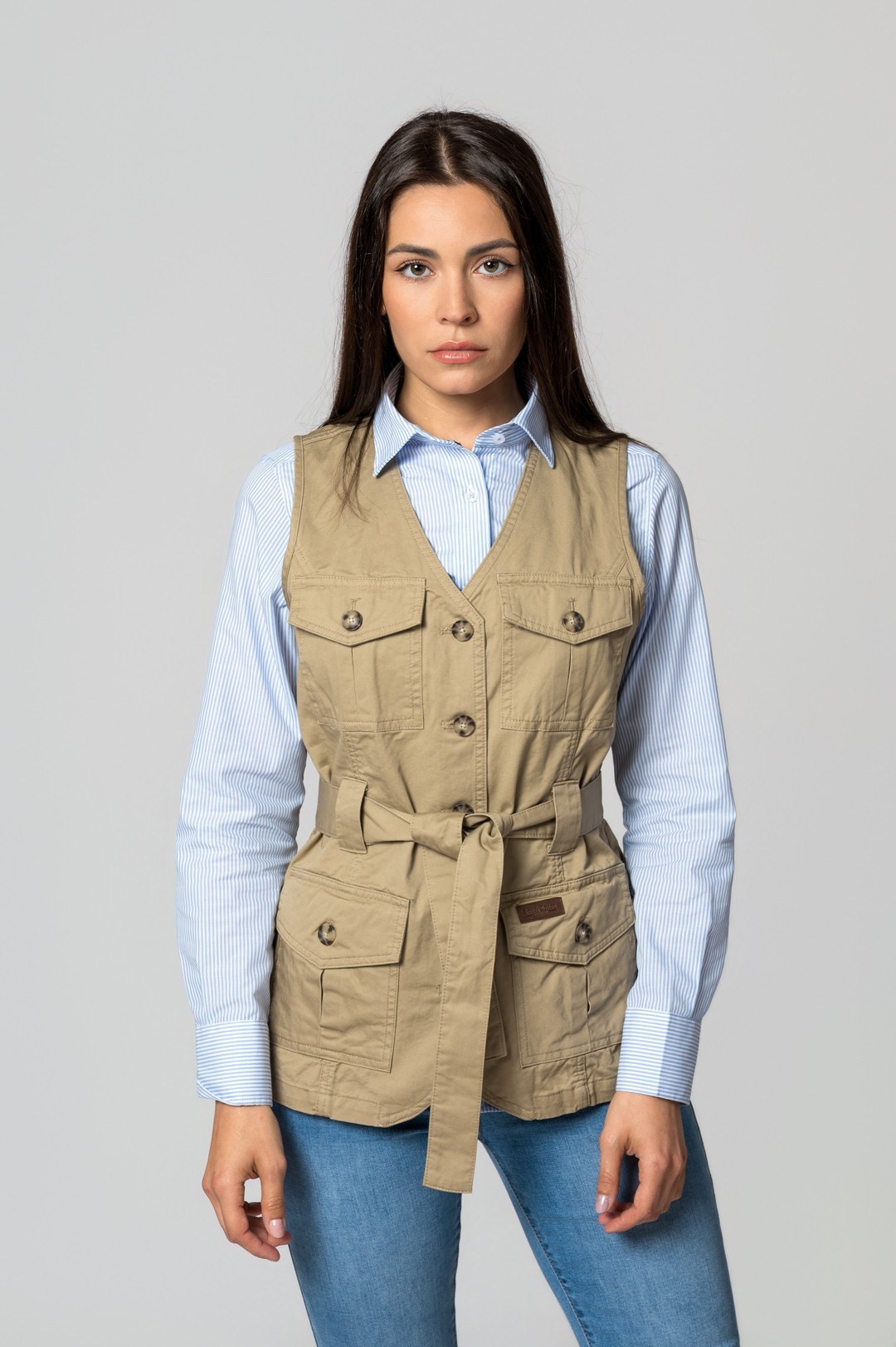 Colete bege com botões e cinto, sobre camisa azul riscada e jeans azuis.