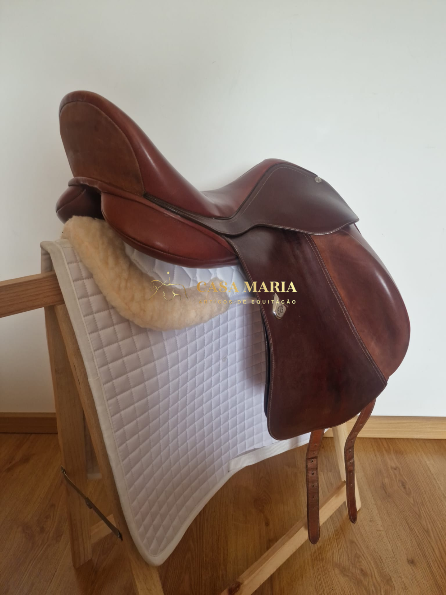 Sela de equitação em couro castanho sobre suporte de madeira com alforge branco