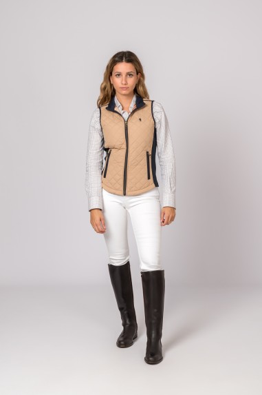 Mulher com colete bege acolchoado, camisa xadrez branca, calças brancas e botas pretas