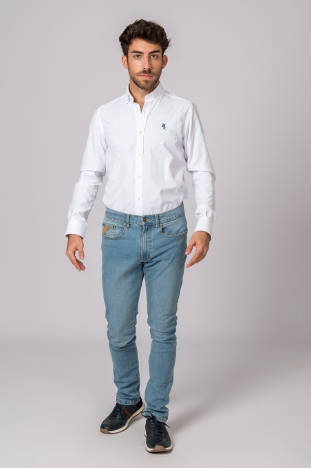 Homem com camisa branca, jeans azul claro e ténis pretos sobre fundo cinzento claro