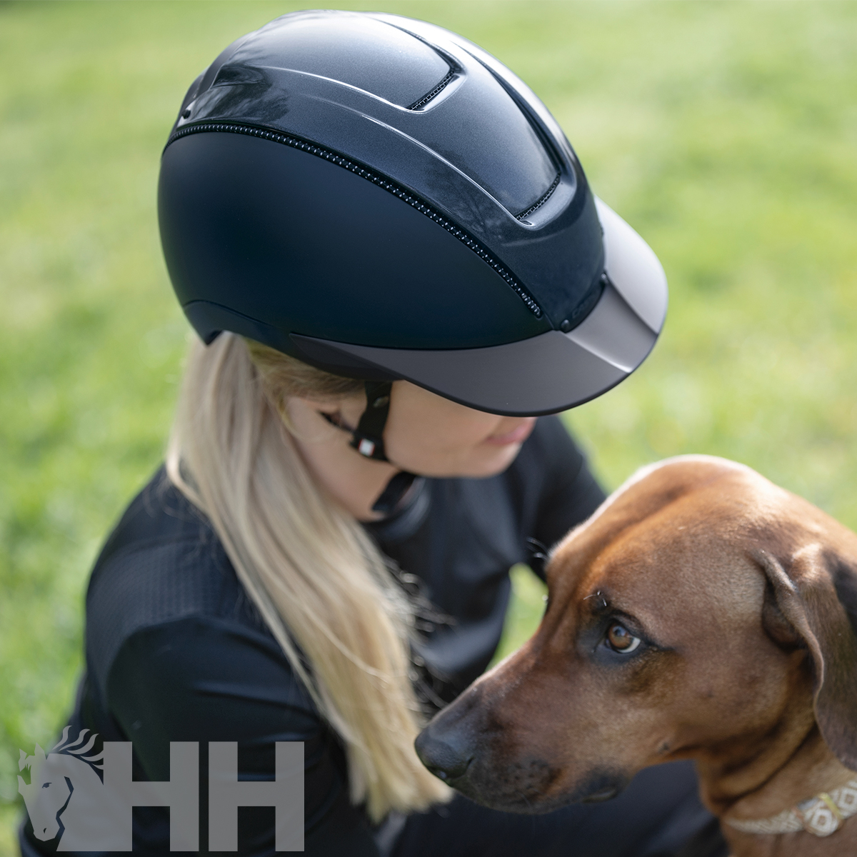 Capacete de equitação preto com detalhes cinza usado por pessoa loira junto a cão castanho