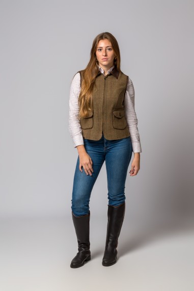 Mulher com colete castanho, camisa às riscas, jeans azuis e botas pretas em fundo cinzento
