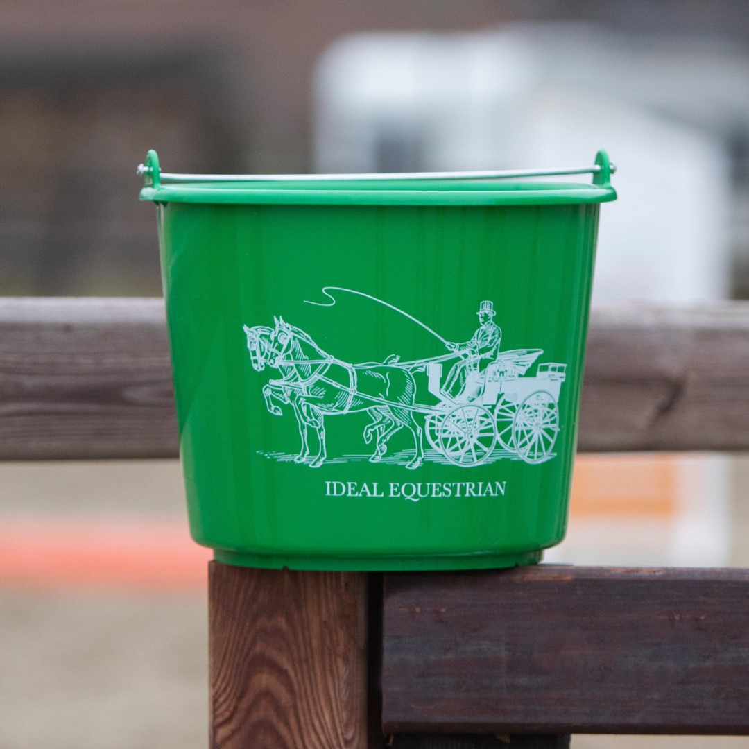 Balde verde de plástico com ilustração de cavalo e cocheiro e texto 'IDEAL EQUESTRIAN' pendurado em vedação de madeira.