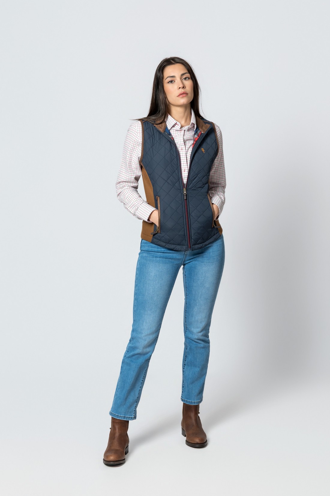 Mulher com colete azul acolchoado, camisa xadrez, jeans e botas castanhas