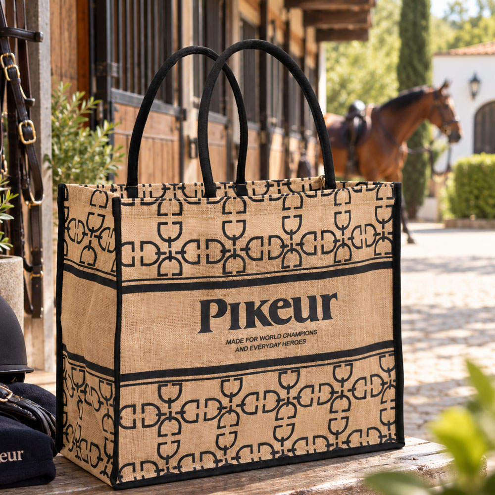 Saco de tecido bege com padrões e texto preto Pikuer num ambiente exterior com cavalo e estábulos ao fundo