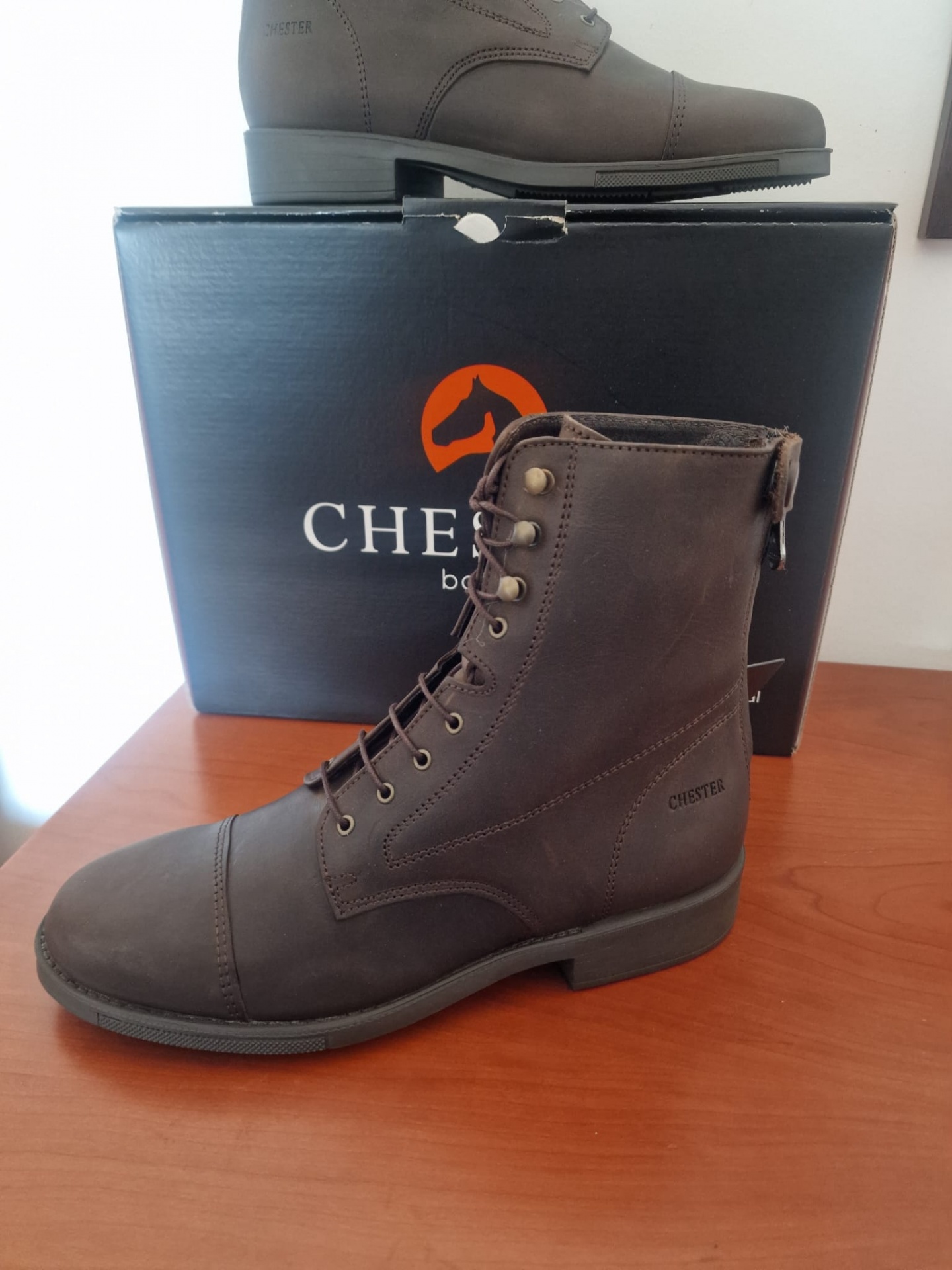 Bota castanha escura em couro com atacadores e sola preta, junto a uma caixa preta com logo CHESTERS