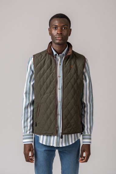 Homem com colete matelassê verde-oliva, camisa às riscas azul e branco, e jeans azuis