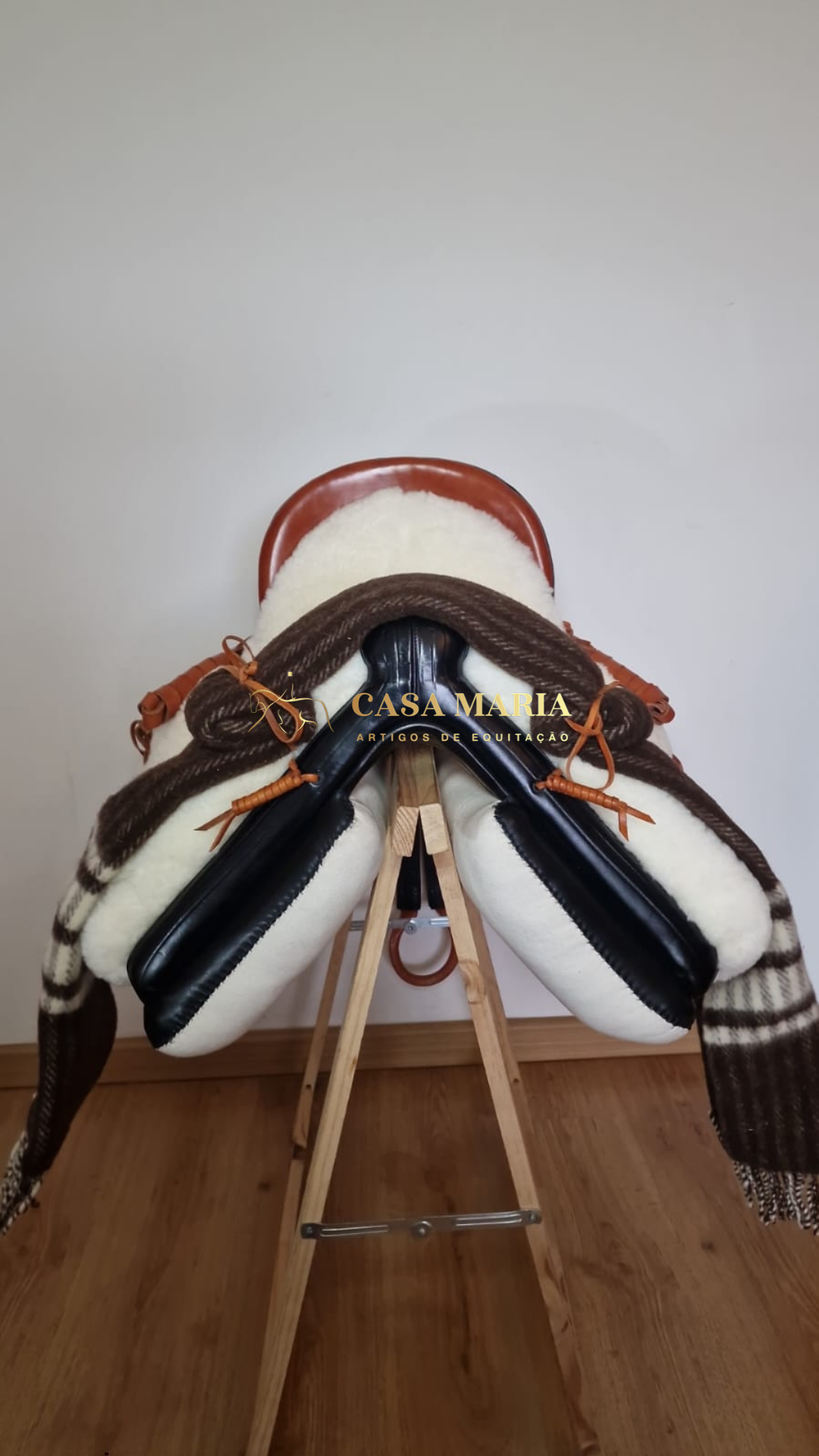 Sela de equitação em couro castanho com acolchoamento branco e manta listrada sobre suporte de madeira