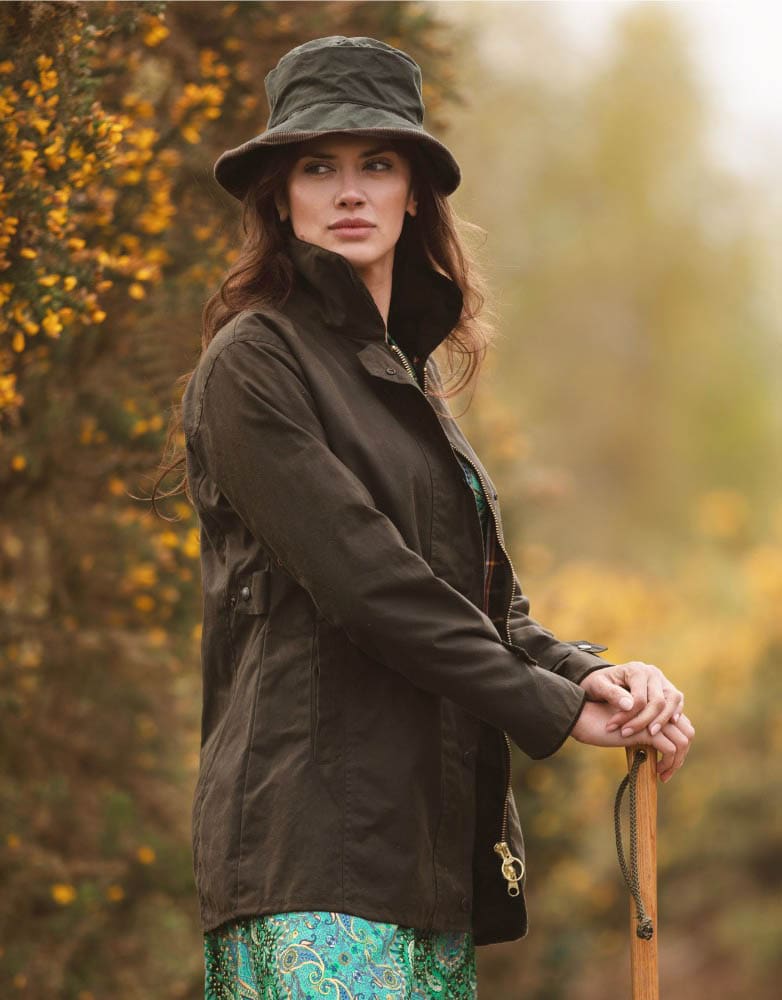 Mulher com chapéu e casaco castanho escuro segurando bastão de madeira na natureza outonal