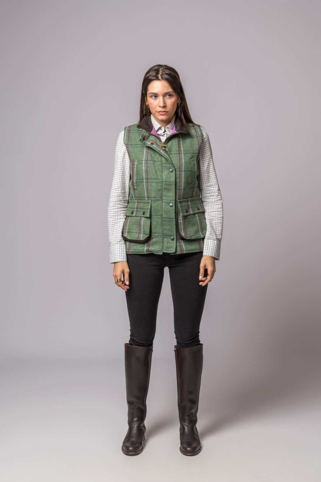 Mulher vestindo colete verde xadrez, camisa branca xadrez, calças pretas e botas castanhas em fundo cinzento