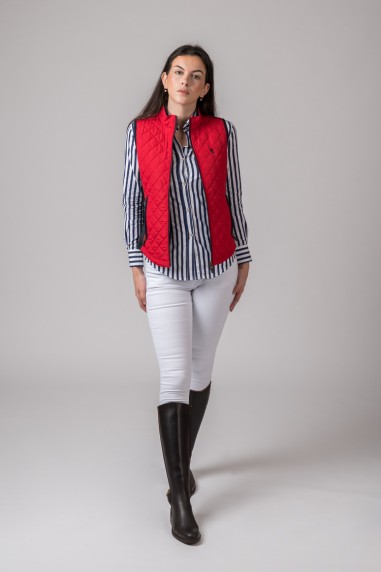 Mulher com colete vermelho, camisa riscada azul e branca, calças brancas e botas pretas em estúdio cinzento