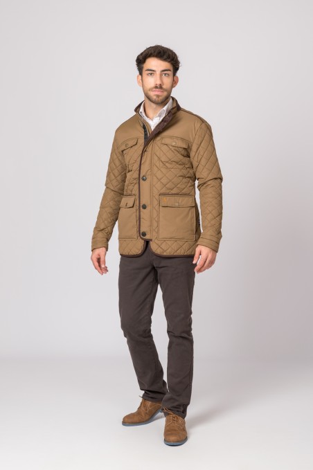 Homem com casaco matelassê castanho, calças castanhas escuras e sapatos castanhos em fundo cinzento.