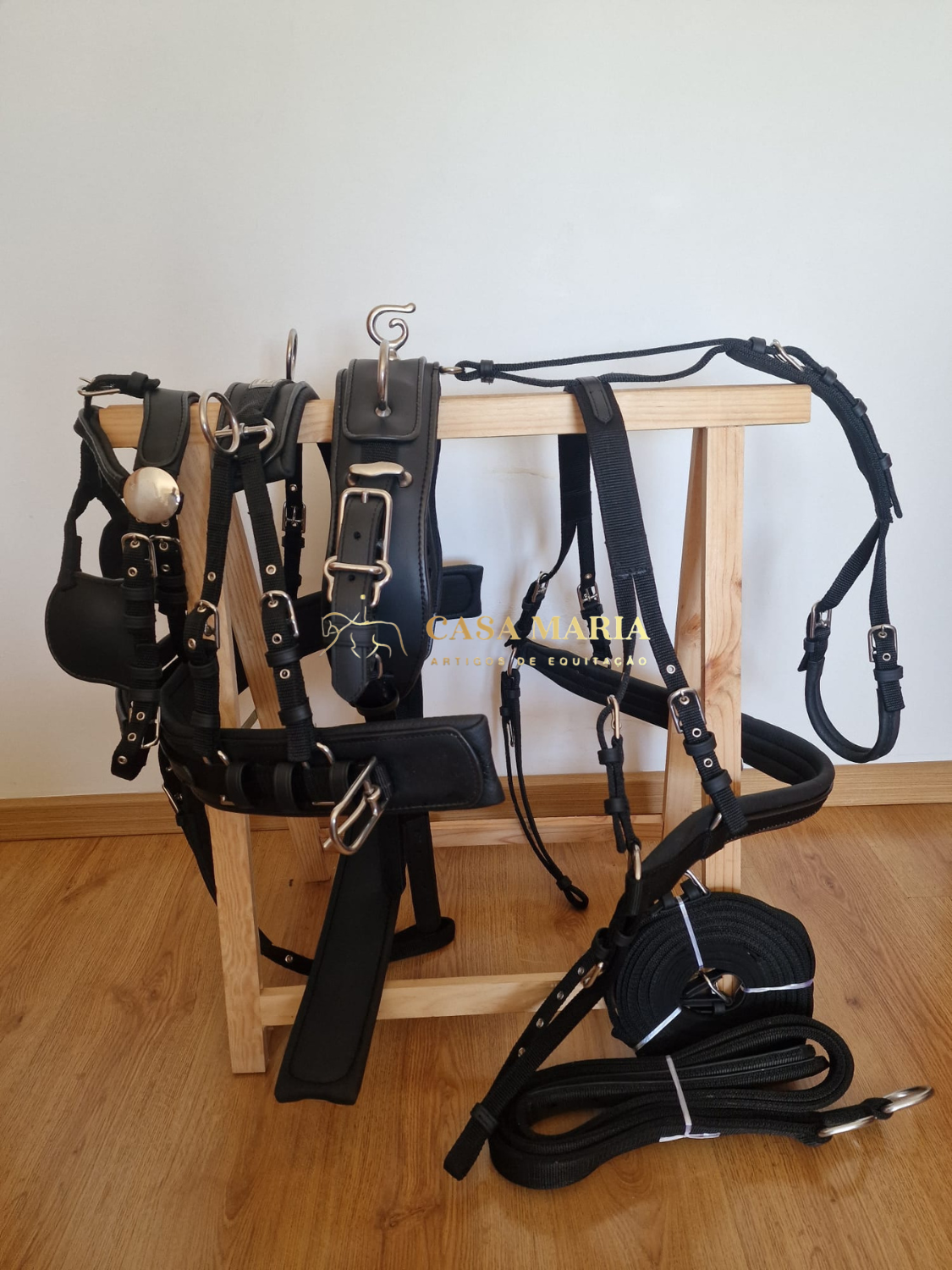 Equipamentos de equitação em couro e nylon preto com fivelas metálicas pendurados em estrutura de madeira.