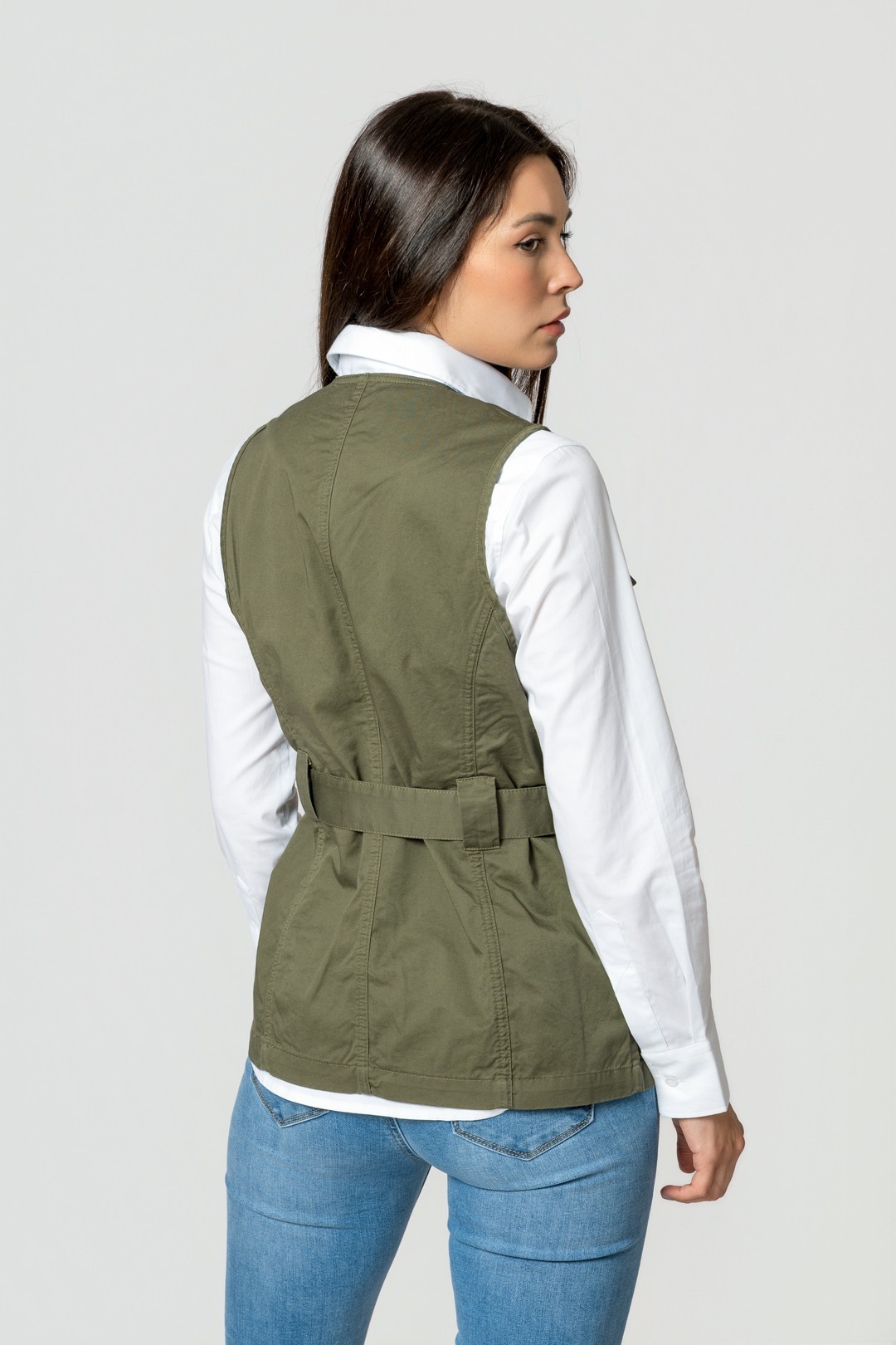 Mulher vista de costas com colete verde caqui, camisa branca e jeans azul