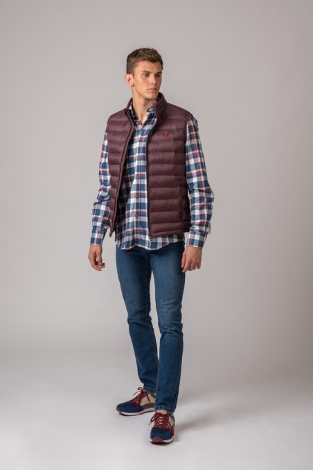 Homem com colete bordeaux, camisa às quadrículas, jeans e sapatilhas em fundo cinza